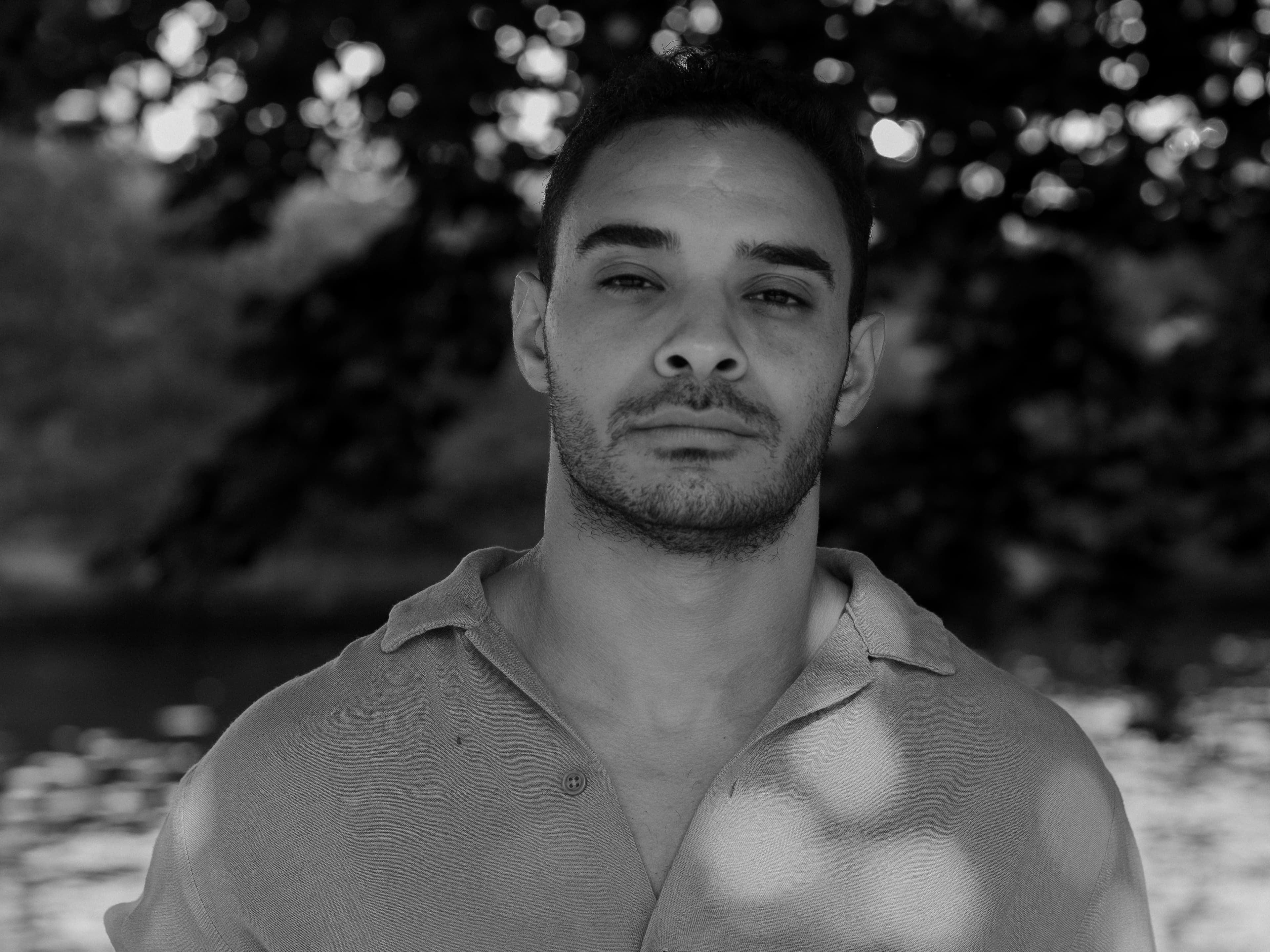 A black and white portrait of Hithem standing by the Canal, with a serene river flowing in the background and looking directly at the camera.